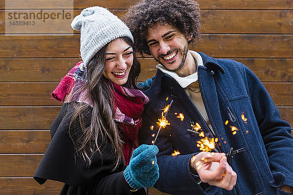 Glückliches junges Paar in Winterkleidung hält Wunderkerzen vor Holzwand