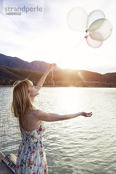 Junge Frau in Sommerkleid mit Blumenmuster steht am Steg und schaut auf Luftballons