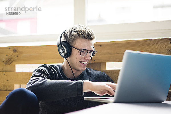 Lächelnder junger Mann mit Laptop  der mit Kopfhörern Musik hört