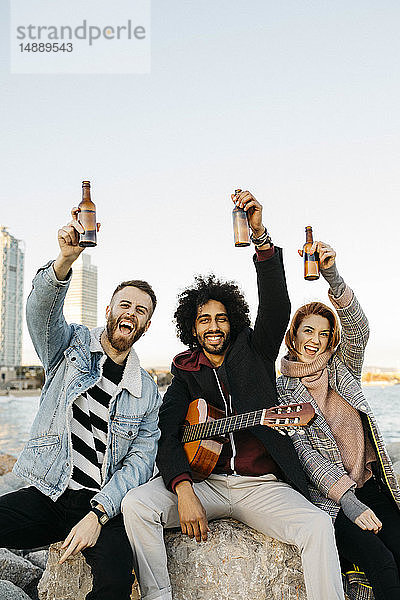 Porträt von drei glücklichen Freunden mit Gitarre  die bei Sonnenuntergang an der Küste Bierflaschen hochheben