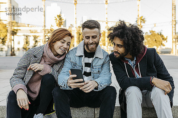 Drei glückliche Freunde sitzen im Freien und schauen auf ihr Handy
