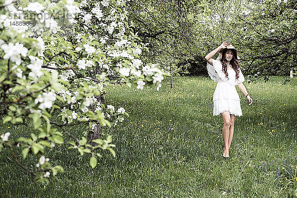 Junge Frau in weißem Kleid und Schlapphut geht barfuß im Garten mit blühenden Apfelbäumen