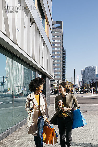 Zwei glückliche Frauen mit Einkaufstaschen und Kaffee zum Mitnehmen spazieren durch die Stadt