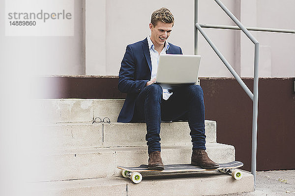 Glücklicher junger Geschäftsmann mit Skateboard  der mit seinem Laptop draußen auf der Treppe sitzt