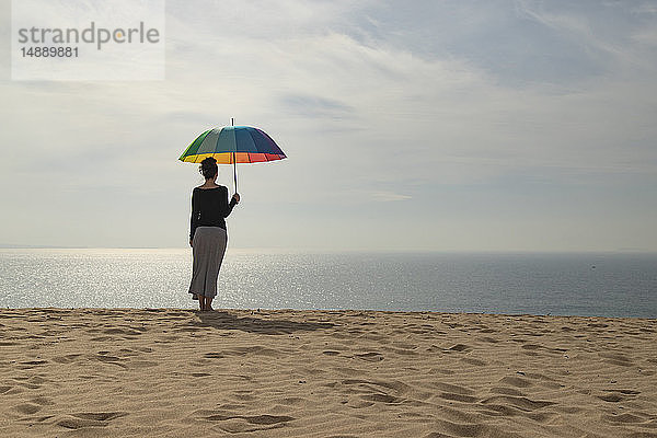 Frau mit buntem Regenschirm am Strand stehend  Rückansicht