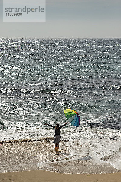 Frau mit buntem Regenschirm am Strand stehend  die Arme erhoben