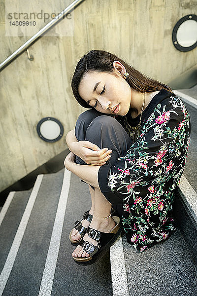 Porträt einer jungen Frau mit geschlossenen Augen auf einer Treppe sitzend und die Knie umarmend