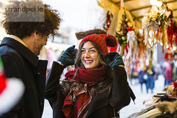 Glückliches junges Paar auf dem Weihnachtsmarkt mit Frau  die einen Wollhut anprobiert