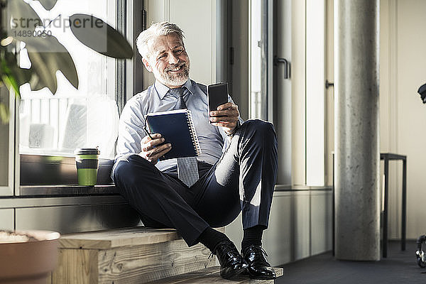 Lächelnder reifer Geschäftsmann am Fenster sitzend mit Handy und Notebook