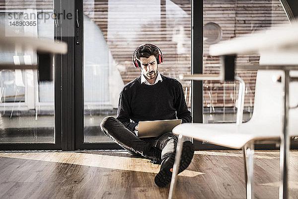 Geschäftsmann sitzt auf dem Boden seines Büros und benutzt Laptop und Kopfhörer