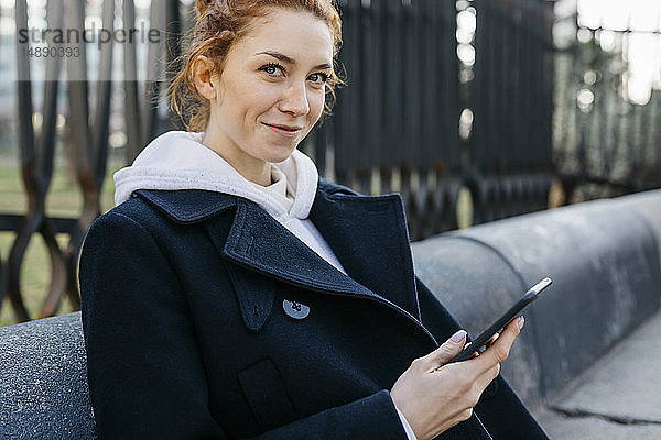 Porträt einer lächelnden jungen Frau  die auf einer Bank sitzt und ein Mobiltelefon hält
