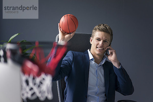 Junger Geschäftsmann sitzt am Schreibtisch im Büro  spielt Basketball und telefoniert