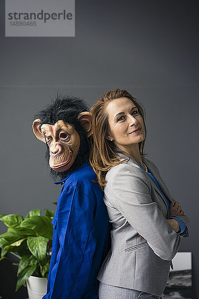 Geschäftsfrau  Rücken an Rücken mit einem Auszubildenden stehend  mit Affenmaske und blauer Evrall