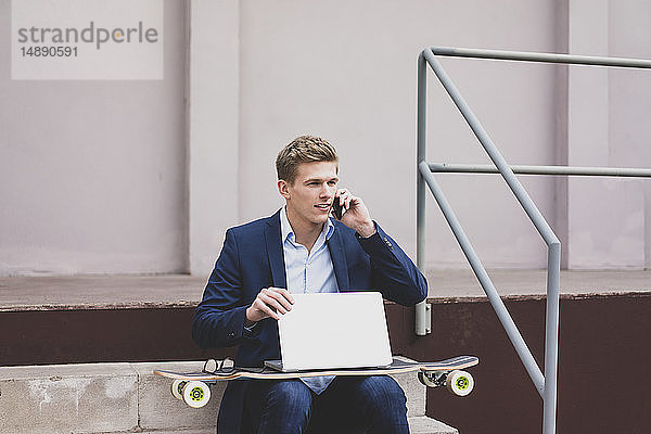 Junger Geschäftsmann mit Skateboard und Laptop sitzt draußen auf der Treppe und telefoniert mit dem Handy
