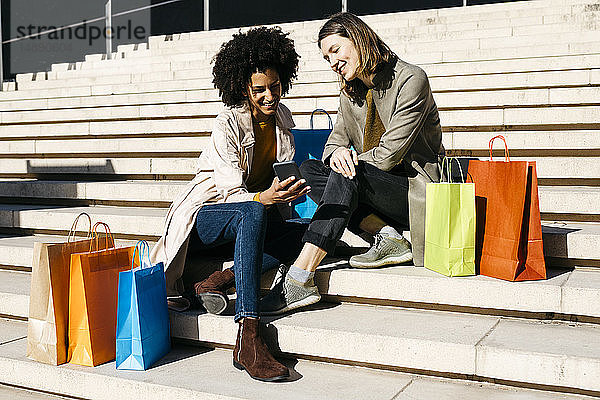 Zwei glückliche Frauen mit Einkaufstaschen sitzen auf der Treppe und benutzen ihr Handy