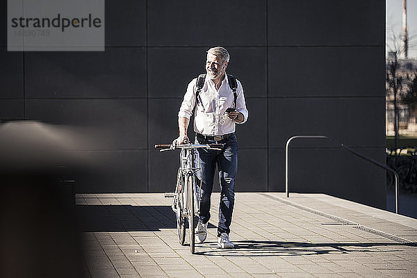 Lächelnder reifer Geschäftsmann mit Fahrrad  Handy und Kopfhörern unterwegs