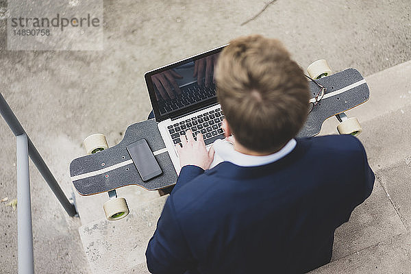 Junger Geschäftsmann mit Skateboard sitzt im Freien auf einer Treppe mit Laptop