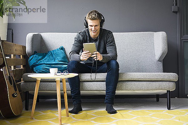 Lächelnder junger Mann mit Gitarre  Tablett und Kopfhörer auf Couch sitzend
