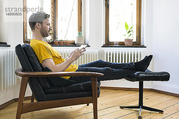 Lässiger Mann in gelbem Hemd sitzt in Lounge Chair in stilvoller Wohnung und schaut auf Tablett