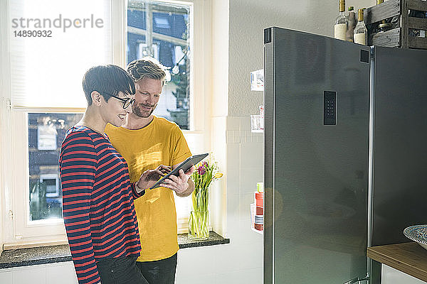 Glückliches Paar mit Tablette vor dem Kühlschrank beim Online-Einkauf von Lebensmitteln