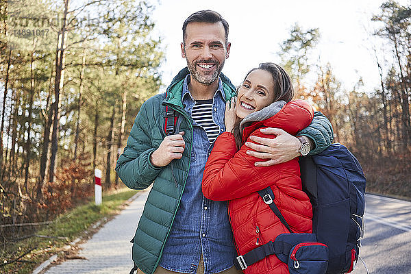 Porträt eines glücklichen Paares  das sich während einer Rucksacktour auf einer Straße im Wald umarmt