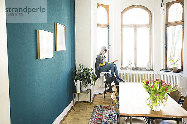 Frau sitzt auf dem Fensterbrett in stilvoller Wohnung  hält Kaffeetasse und liest ein Buch