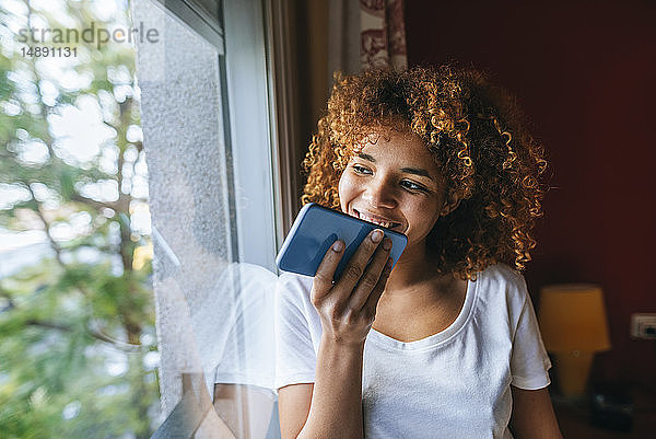 Junge Frau mit lockigem Haar sendet Sprachnachricht mit Handy neben dem Fenster