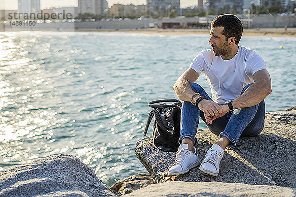 Spanien  Barcelona  Mann sitzt auf einem Felsen vor dem Meer und schaut in die Ferne