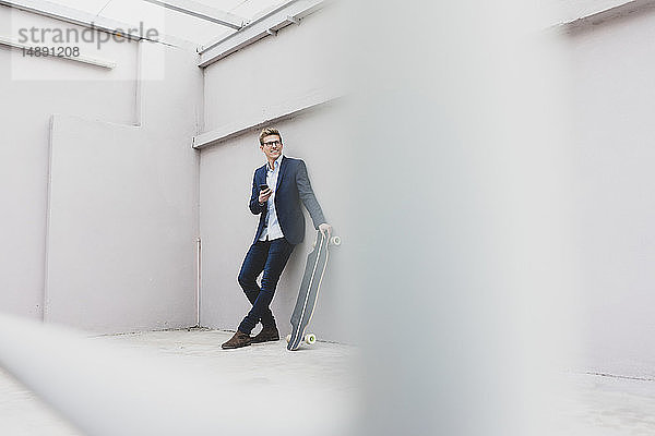 Lächelnder junger Geschäftsmann mit Skateboard und Handy an einer Betonwand lehnend