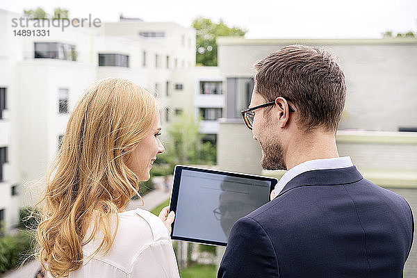 Immobilienmakler steht mit einem Kunden auf einem Balkon und schaut auf ein digitales Tablett