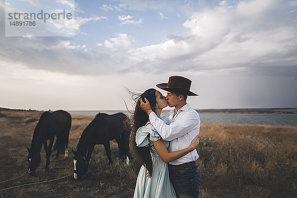 Junges Paar küsst sich auf einem Feld bei Pferden