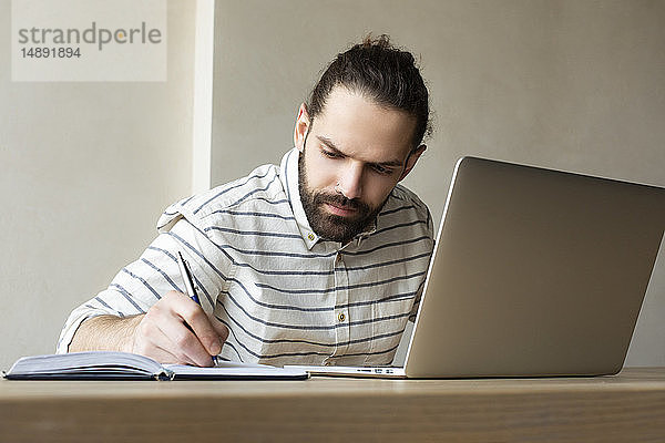 Junger Geschäftsmann mit Laptop  der in einen Notizblock schreibt