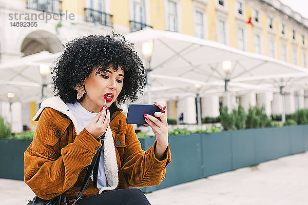 Junge Frau trägt mit ihrem Smartphone roten Lippenstift auf