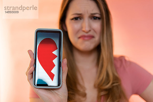 Junge Frau hält Smartphone und zeigt gebrochenes Herz