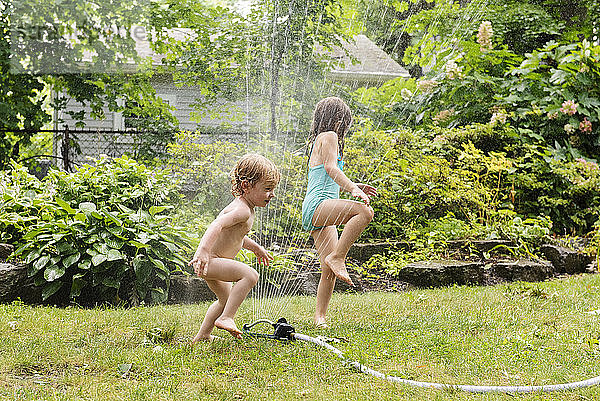 Nackter Junge und seine Schwester spielen mit Sprinkleranlage