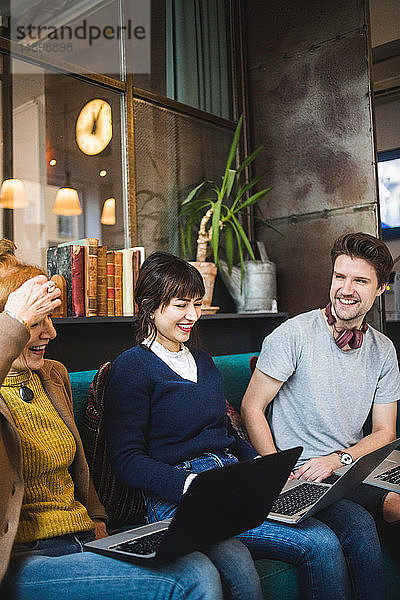 Lächelnde männliche und weibliche Kollegen schauen auf die Managerin  die mit Laptop auf dem Sofa im Kreativbüro sitzt