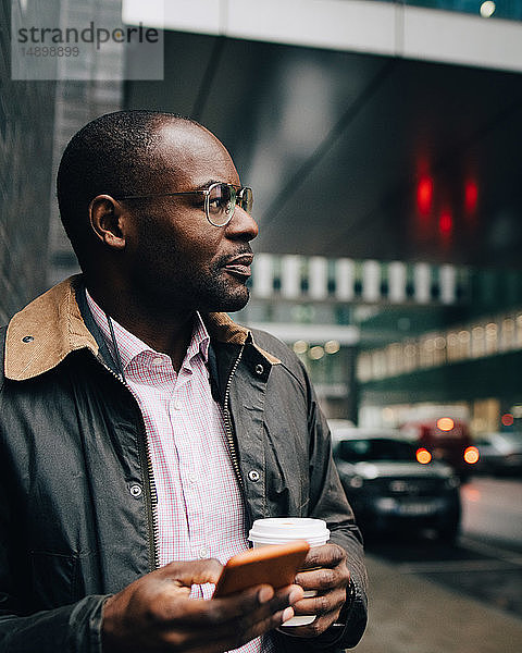 Geschäftsmann hält Kaffee und Mobiltelefon in der Hand und schaut weg  während er in der Stadt auf der Straße steht