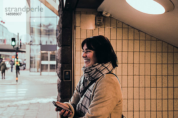 Fröhliche junge Frau in warmer Kleidung  die im Winter am Eingang der U-Bahn in der Stadt steht