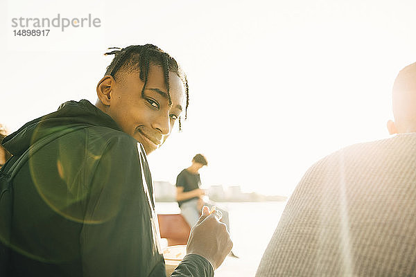 Porträt eines lächelnden Teenagers  der eine Mahlzeit hält  während er mit einem Freund an einem sonnigen Tag auf der Promenade sitzt
