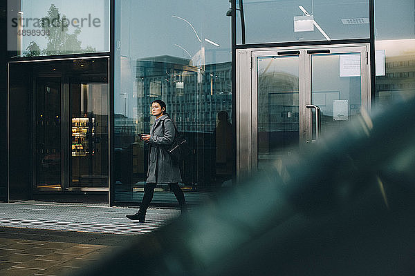 Geschäftsfrau mit Smartphone schaut weg  während sie auf dem Bürgersteig gegen ein Gebäude in der Stadt läuft