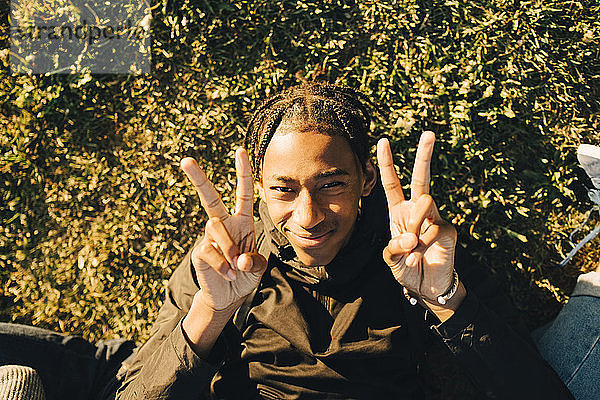 Hochwinkel-Porträt eines lächelnden Teenagers  der an einem sonnigen Tag ein Friedenszeichen gegen Pflanzen gestikuliert