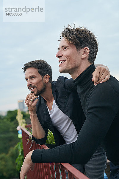 Lächelnde männliche Freunde schauen weg  während sie am Geländer auf der Terrasse gegen den Himmel stehen