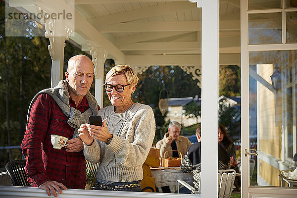 Fröhliche reife Frau zeigt kahlköpfigem Mann mit Freunden im Hintergrund auf der Veranda ihr Handy