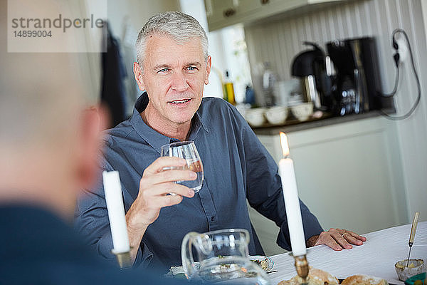 Reifer Mann schaut weg  während er ein Trinkglas auf dem Esstisch hält  mit einem Freund im Vordergrund
