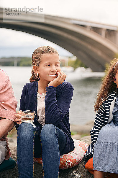 Lächelndes Mädchen schaut weg  während es am Seeufer mit der Familie im Park beim Picknick ein Getränk hält