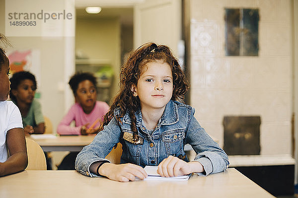 Porträt eines lächelnden Schulmädchens  das mit Freunden am Schreibtisch im Klassenzimmer sitzt
