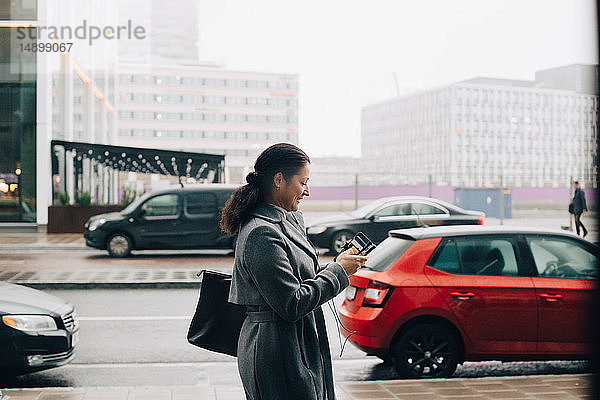 Seitenansicht einer Geschäftsfrau  die ein Smartphone benutzt  während sie auf dem Bürgersteig gegen den Himmel in der Stadt läuft