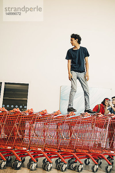 Sorgloser Teenager steht auf gestapelten roten Einkaufswagen vor dem Einkaufszentrum