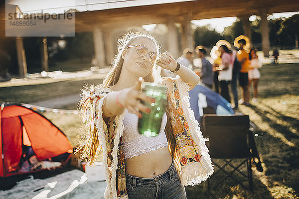 Porträt einer verspielten jungen Frau  die ein Getränk in der Hand hält  während sie auf ein Ereignis an einem sonnigen Tag zeigt