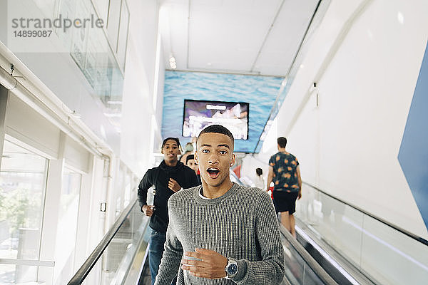 Porträt eines schockierten Teenagers  der sich mit einem Freund im Hintergrund auf der Rolltreppe im Einkaufszentrum nach unten bewegt